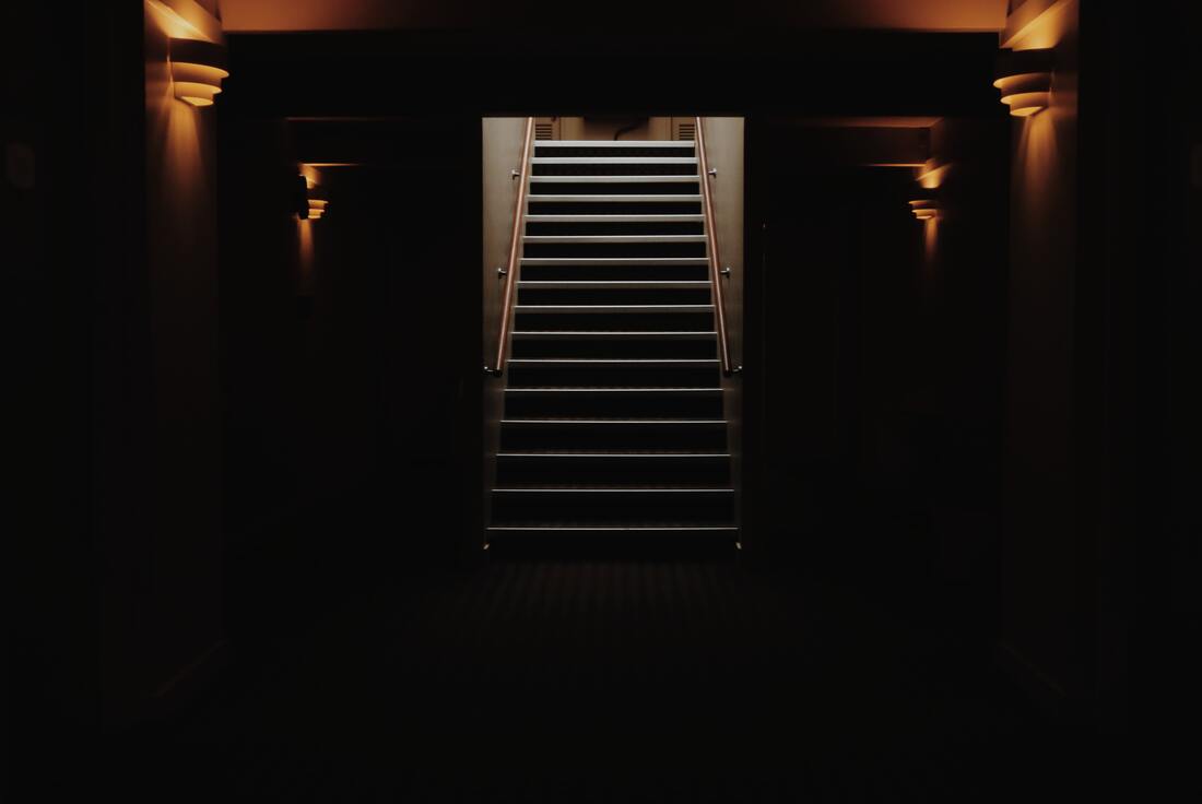 A photo from the bottom of a staircase in a dimly lit basement. The light at the top of the stairs barely illuminates the upstairs landing.